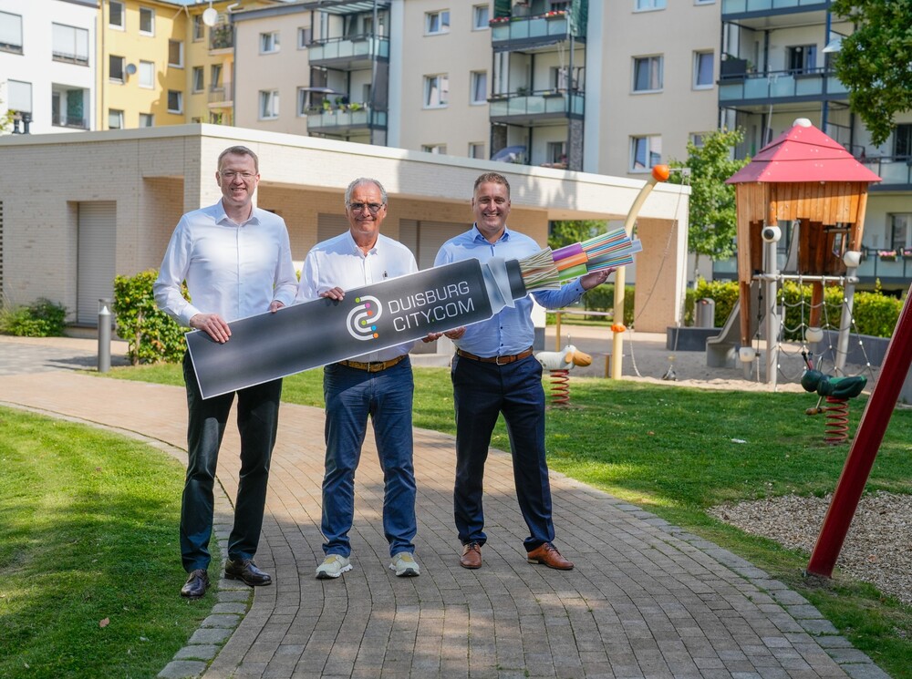 Drei Männer stehen in einem Innenhof mit Spielplatz und Wiesen und halten ein Pappschild hoch, das ein Glasfaserkabel zeigt.