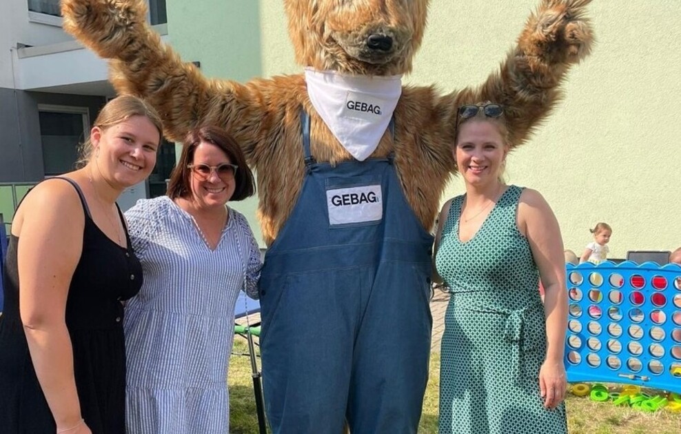 Drei Frauen posieren mit einem Erdmännchen-Maskottchen vor einem Wohnhaus.