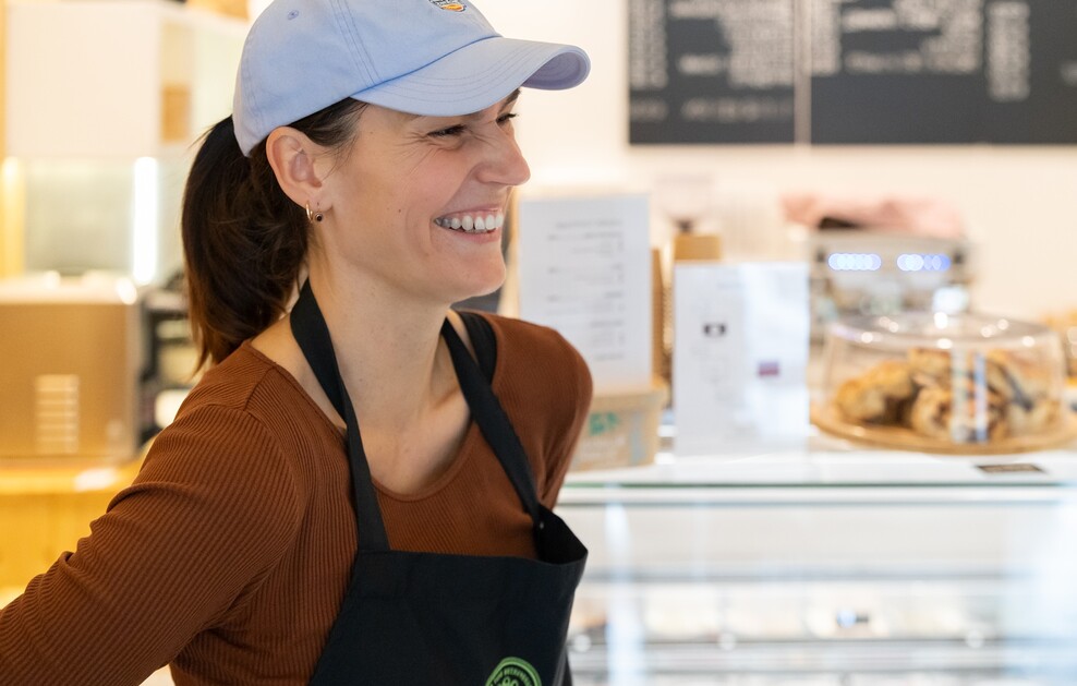 Eine lächelnde junge Frau mit hellblauem Basecap und schwarzer Schürze steht vor einer Theke in einem Eiscafé.