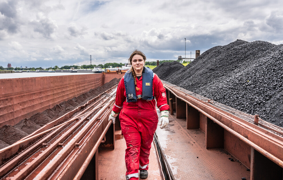 Eine junge Frau in rotem Overall und Schwimmweste geht auf einem fahrenden Kohlefrachter.