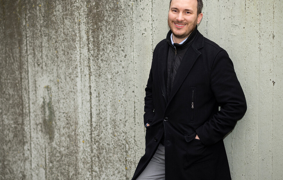 Ein Mann steht lächelnd vor einer Betonwand. Er trägt einen dunklen Mantel und hat die Hände in den Taschen.