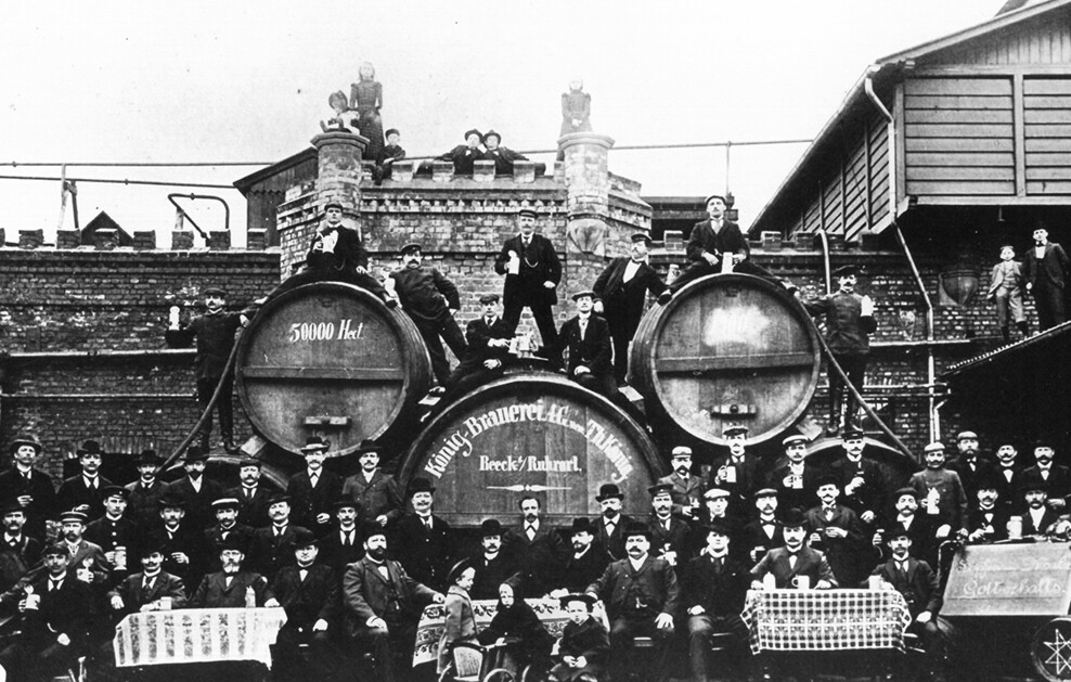 Eine alte Schwarz-Weiß-Fotografie zeigt viele Männer sowie einige Frauen und Kindern vor Bierfässern einer Brauerei