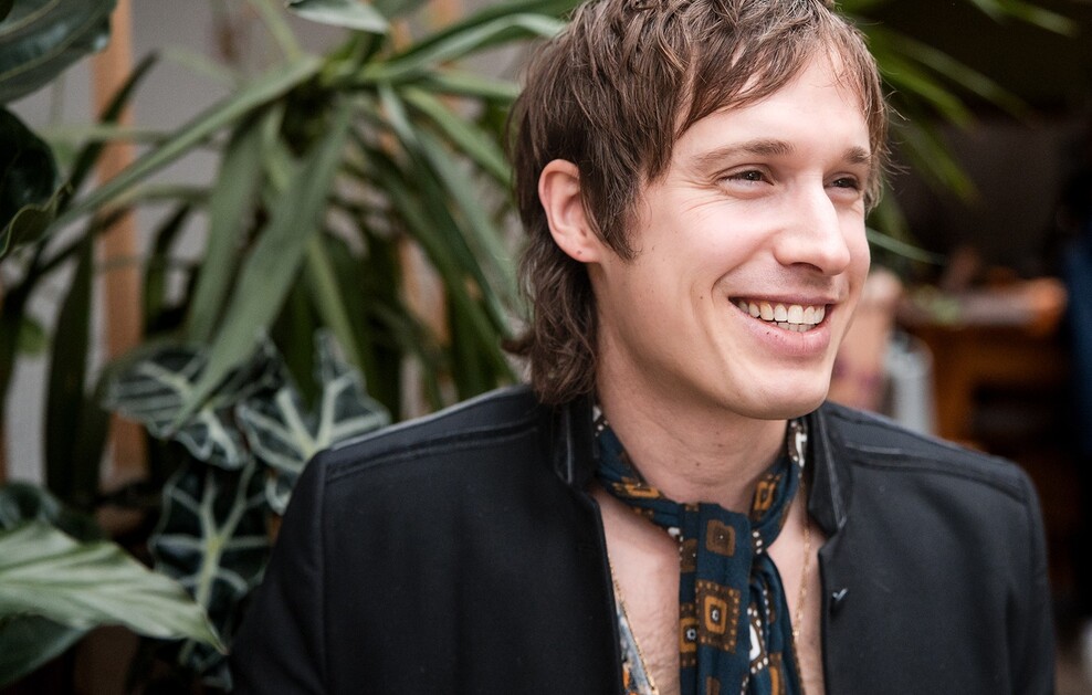 Ein lächelnder junger Mann mit braunen Haaren, bekleidet mit einem schwarzen Sakko und einem gemusterten Schal.