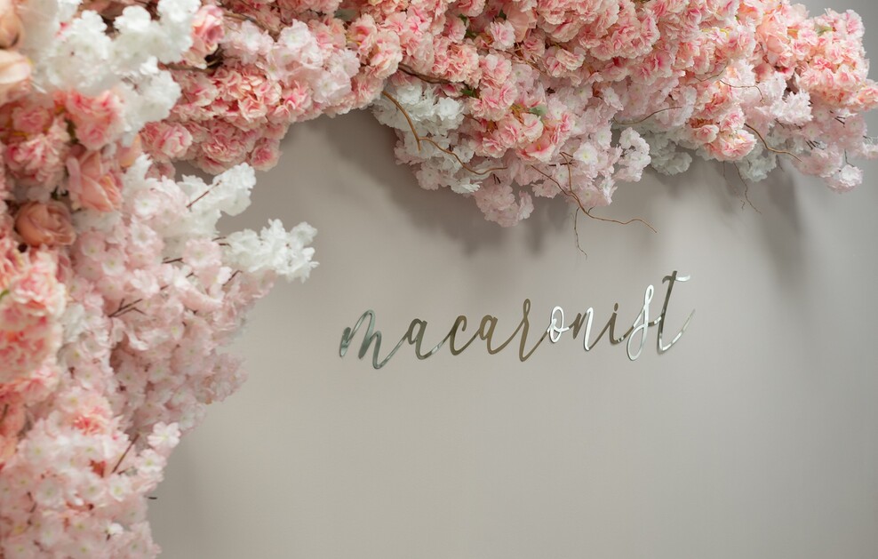 Rosa und weiße Blütenranken schmücken eine helle Wand mit dem Wort "Macaronist" in silbernen Buchstaben.